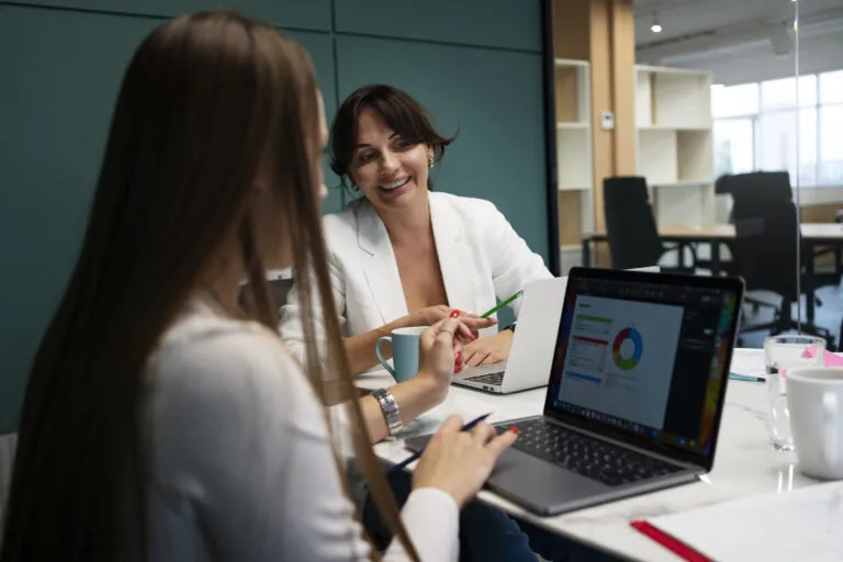 Duas profissionais de marketing analisam gráficos em um notebook durante reunião, discutindo estratégias com IA para clínicas médicas e odontológicas a fim de aumentar agendamentos e reduzir no-show.
